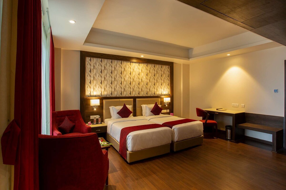A hotel room with a double bed, two bedside tables with lamps, a desk with a chair, and a red curtain.