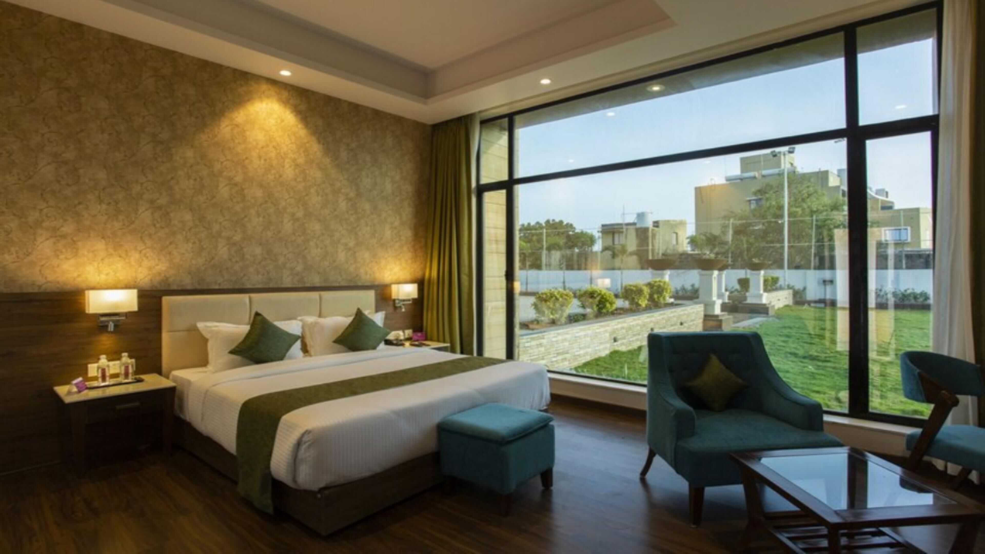A hotel room with a large bed, two bedside tables with lamps, a green upholstered chair, a glass coffee table, and a view of a courtyard through large windows.