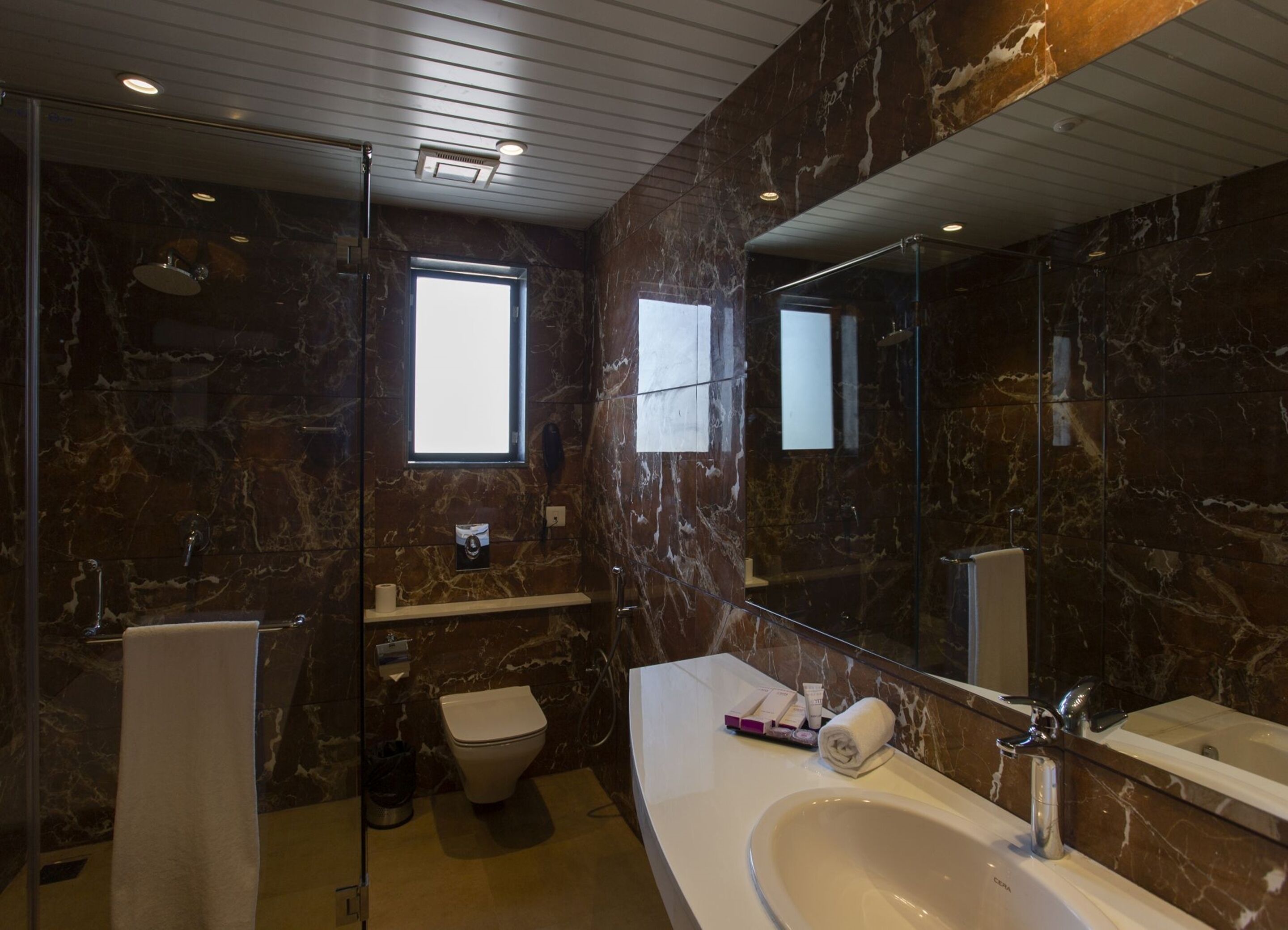 A modern bathroom with a marble wall, a large mirror, a bathtub, and a toilet.