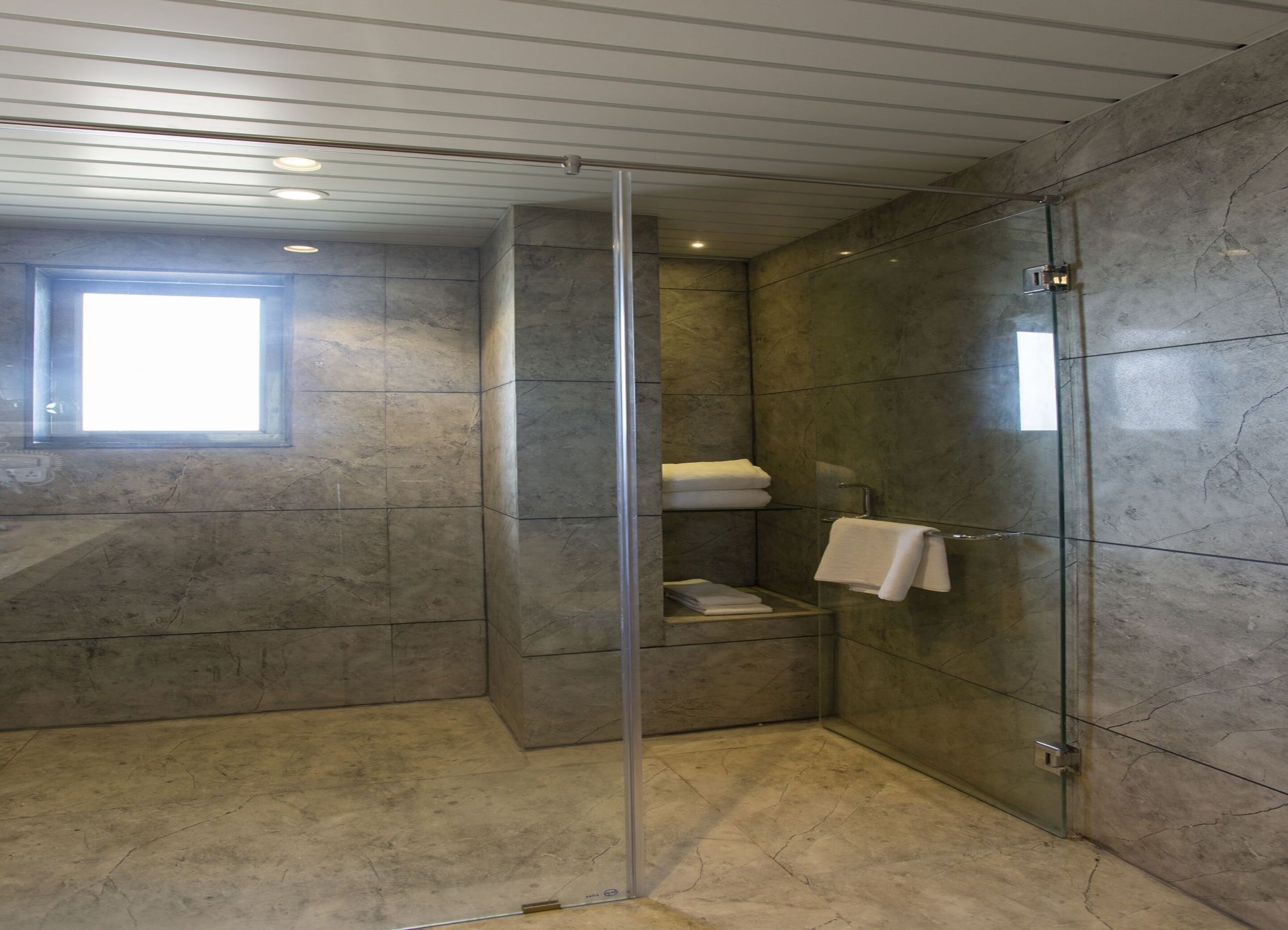 Un baño moderno con cabina de ducha de vidrio, paredes de mármol y un estante empotrado con toallas.