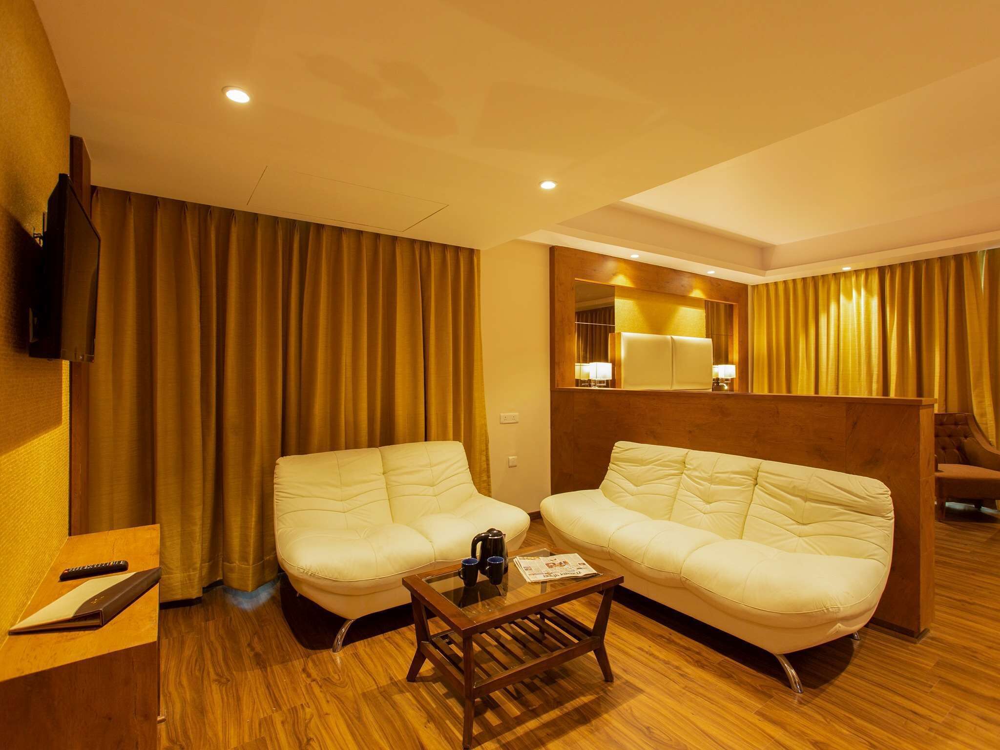 A hotel room with two cream-colored sofas, a wooden coffee table, a TV mounted on the wall, and a wooden desk with a remote control.