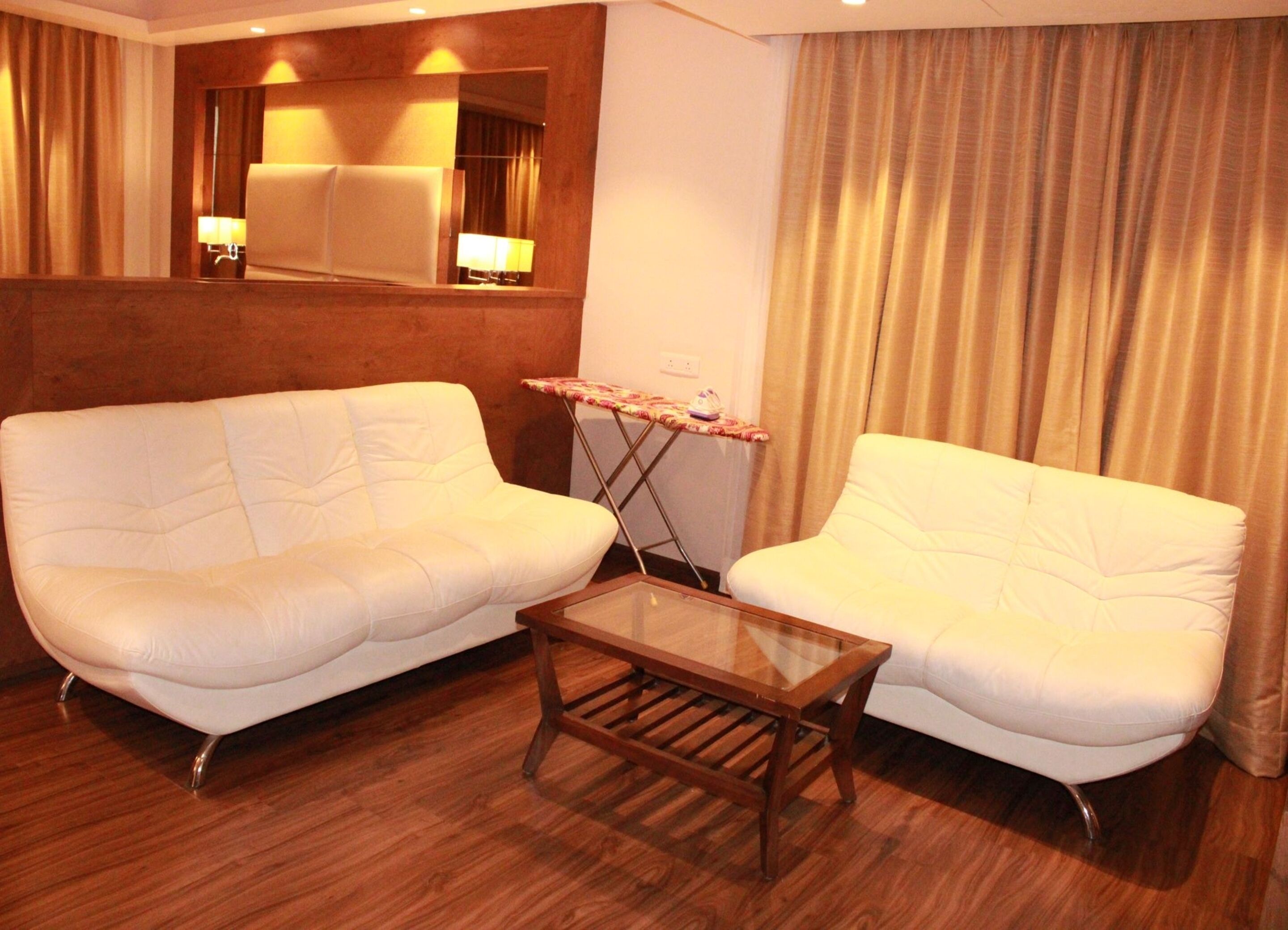 A hotel room with two white sofas, a small wooden coffee table, and a mirror above a wooden partition.