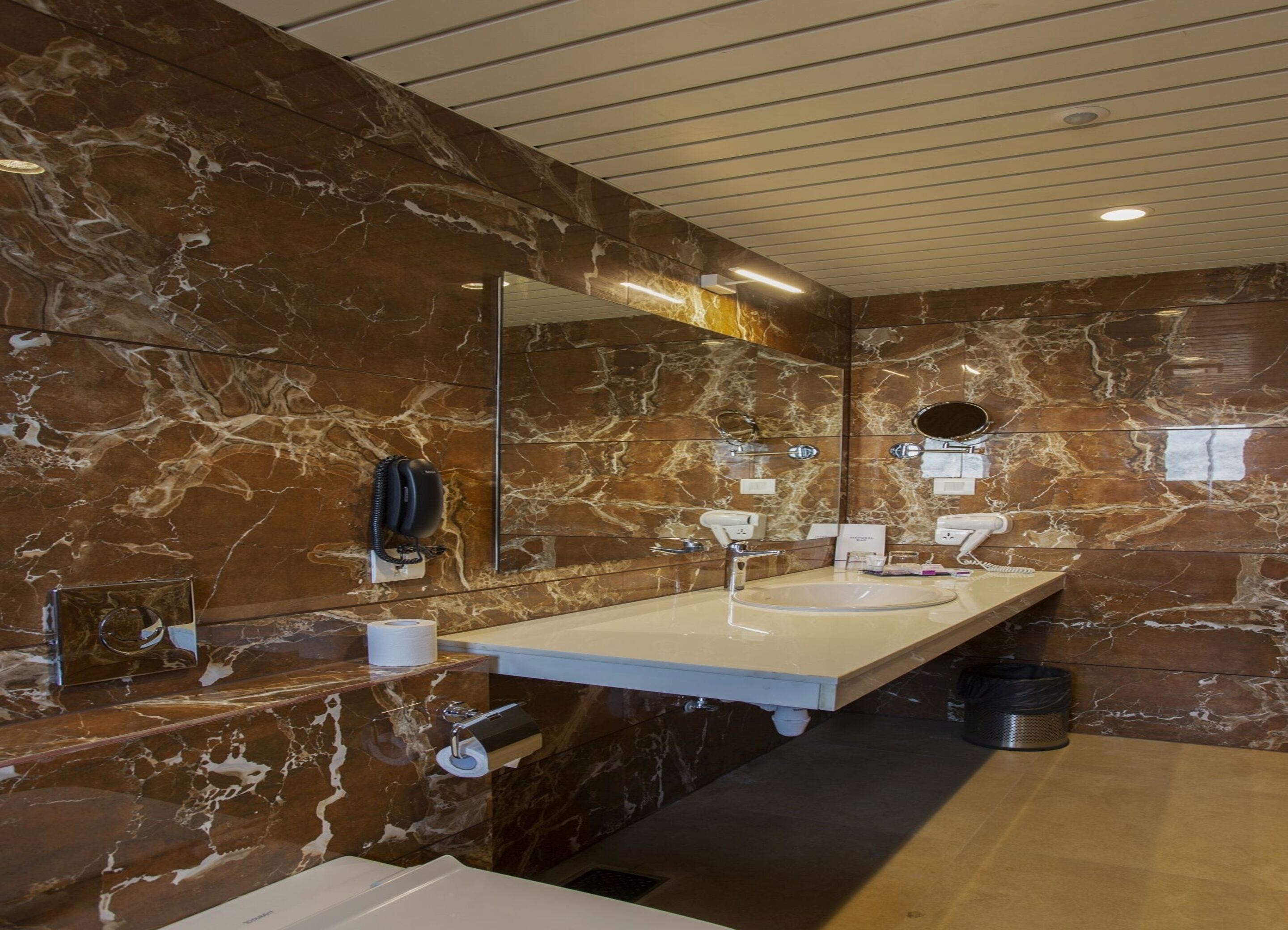 A modern bathroom with marble walls, two sinks, a large mirror, and a phone mounted on the wall.