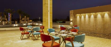 An outdoor dining area with round tables and chairs, a tall pillar, and a decorative wall.