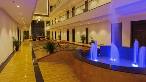 A hotel lobby with a central fountain, multiple floors, and a modern design. - Sarovar Portico Somnath (Veraval)