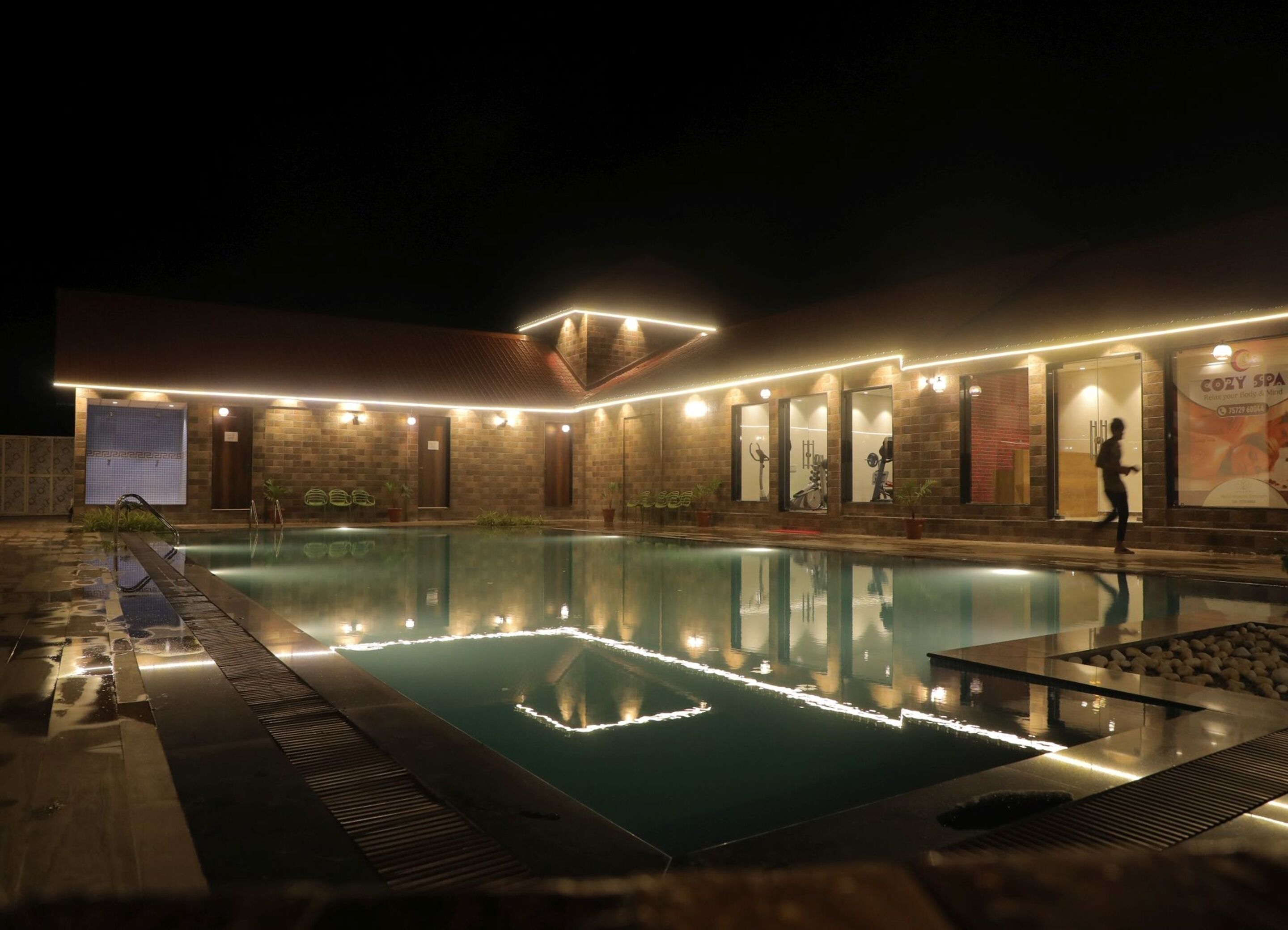 A nighttime view of a building with a swimming pool, illuminated by lights along the roof and poolside.