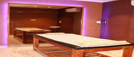 A room with two wooden massage tables, a mirror, a sink, and purple lighting.