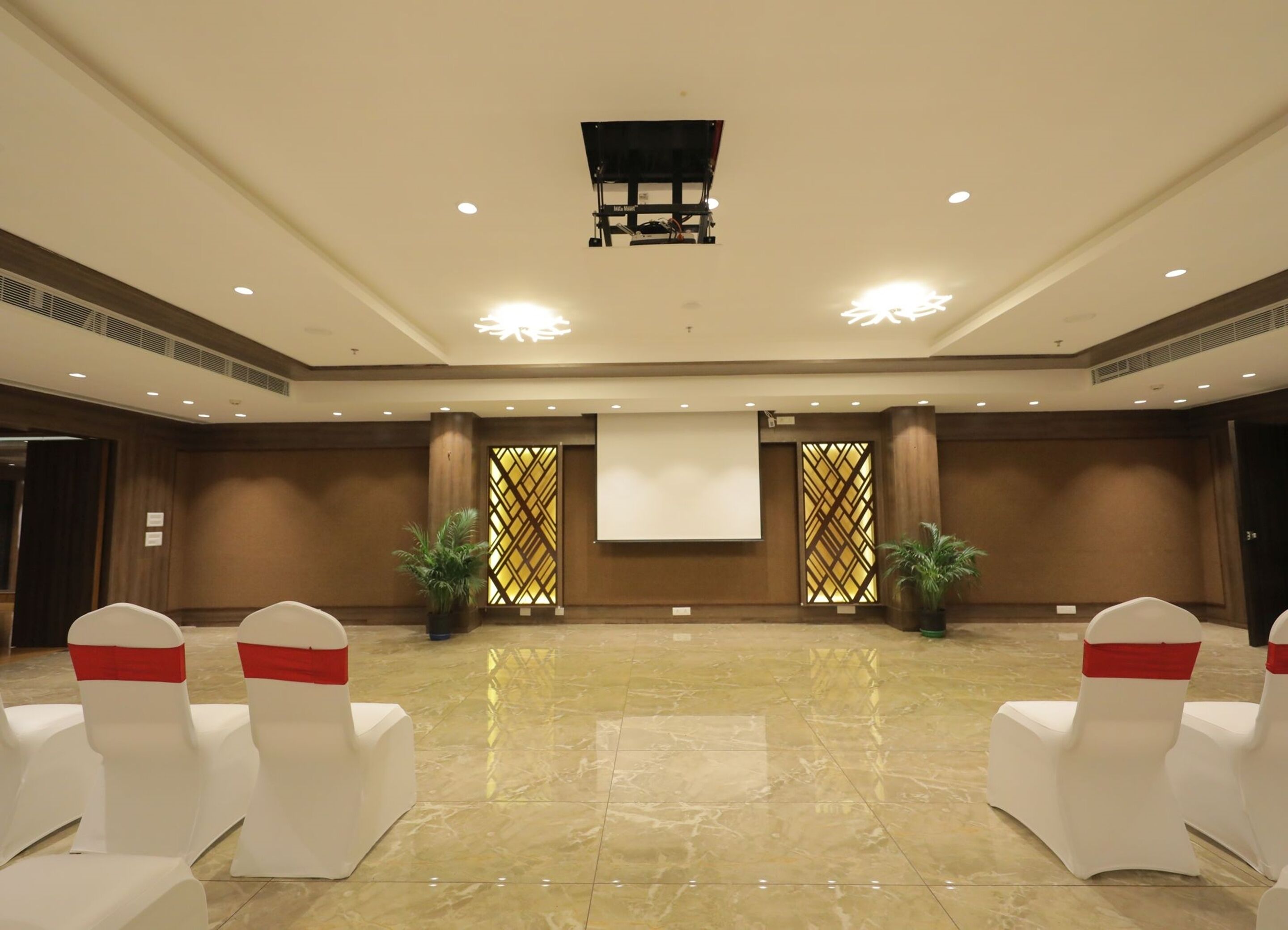 A conference room with a large screen, two potted plants, and white chairs with red sashes.