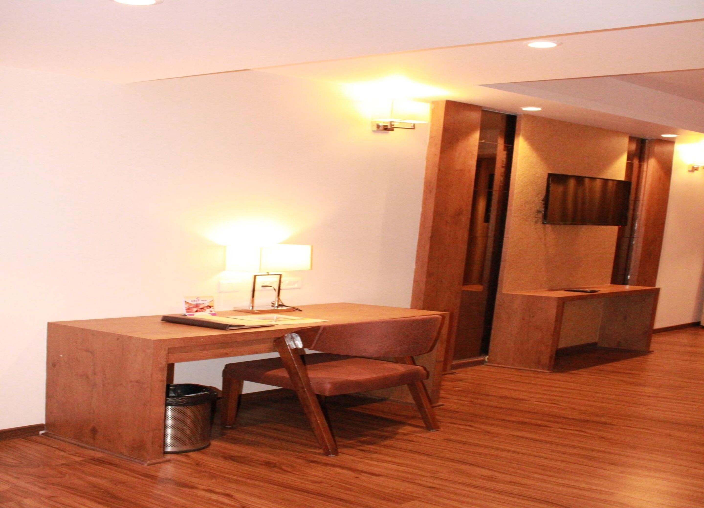 A hotel room with a wooden desk, a chair, a trash bin, and a wall-mounted TV.