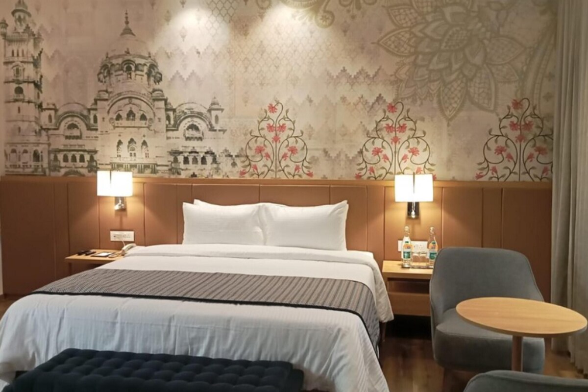 A hotel room with a large bed, two bedside lamps, a small table, a chair, and a decorative wall with architectural designs.