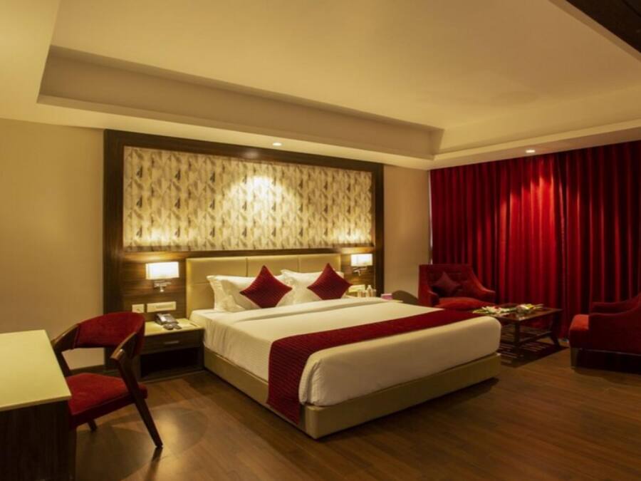 A hotel room with a large bed, a desk, two chairs, a nightstand, and a red curtain.