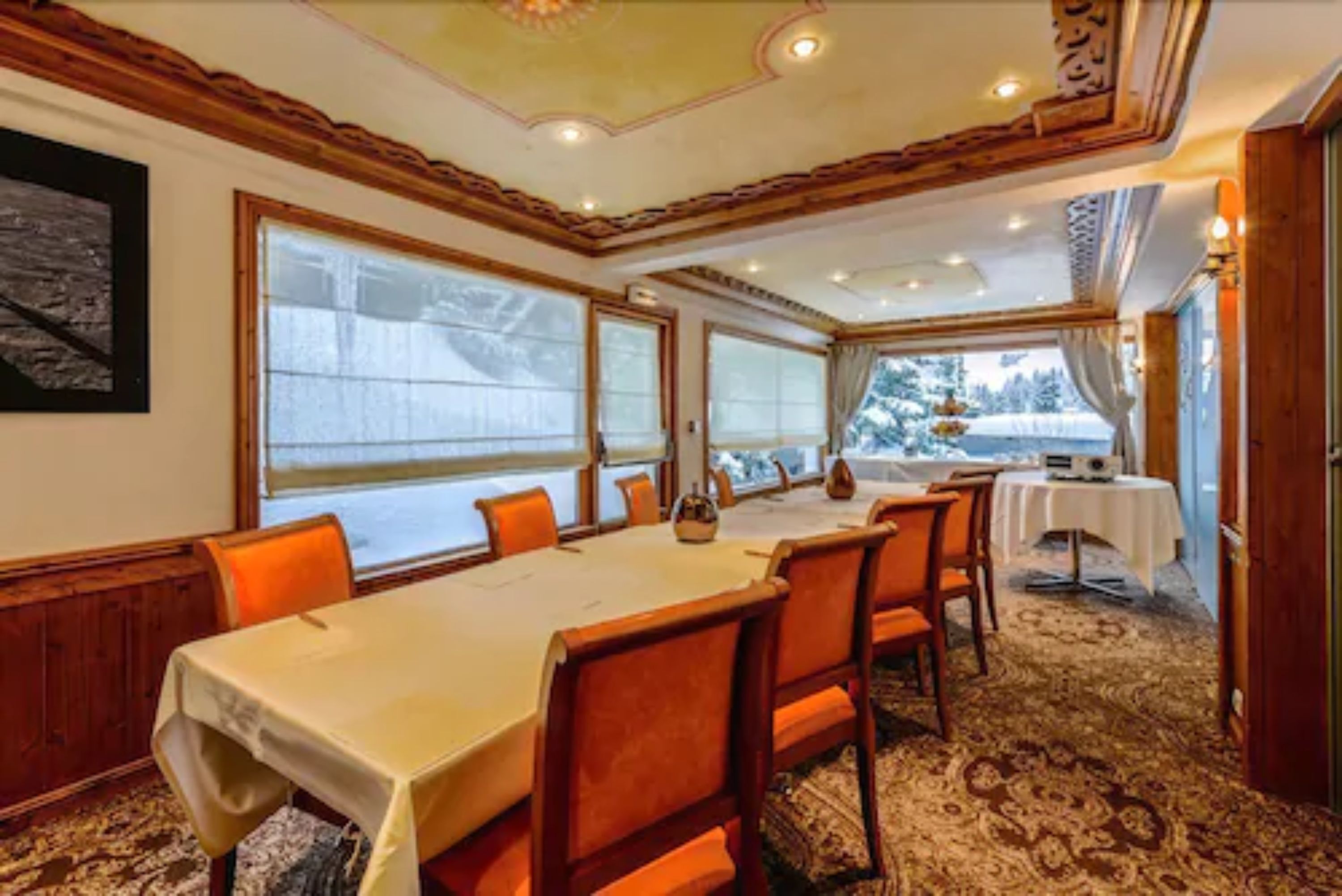 A dining room with a long table, orange chairs, and large windows with a view of trees.