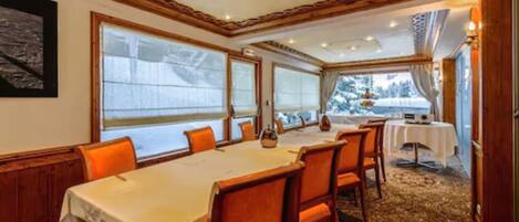 A dining room with a long table, orange chairs, and large windows with a view of trees.