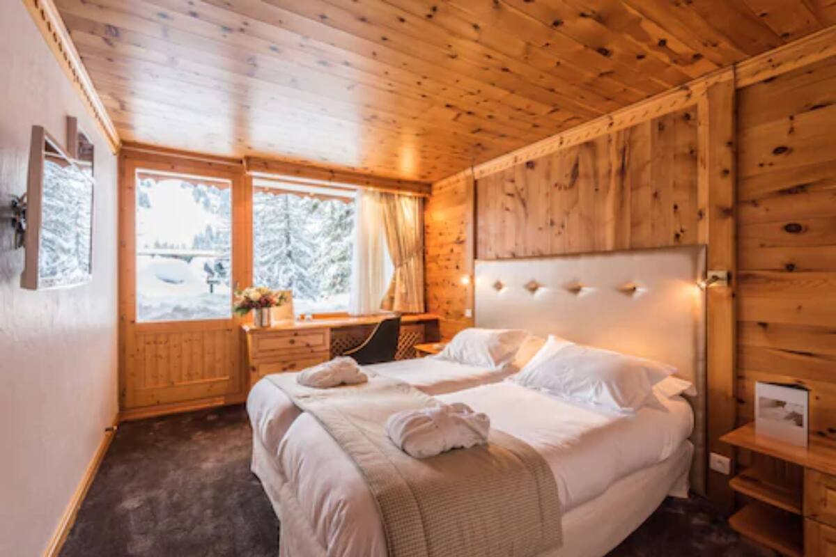 A wooden cabin room with a bed, a desk, and a window with curtains.