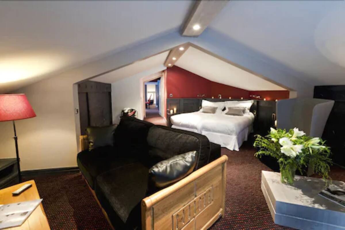 A hotel room with a bed, a black leather sofa, a wooden chair, a small table with a lamp, and a vase with flowers.