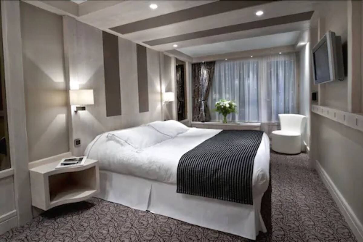 A modern hotel room with a large bed, bedside tables, a TV, and a view of the city.