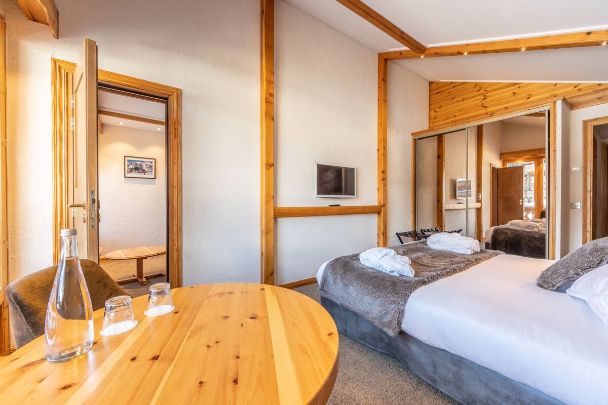 A hotel room with a wooden ceiling, a bed with a gray blanket, a TV on the wall, and a wooden table with a bottle and glasses.
