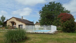 Una casa con piscina, rodeada por una cerca blanca y un gran árbol de hojas rojas.