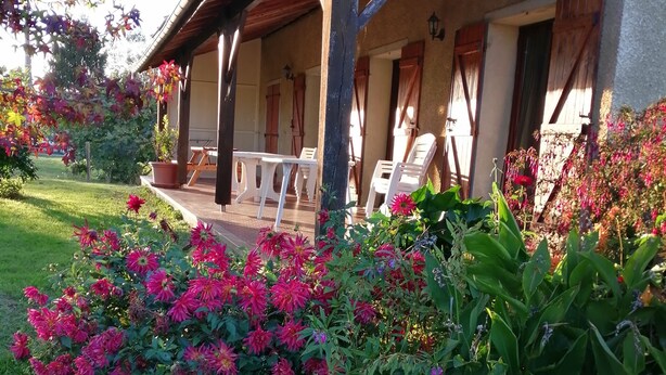 Un área de estar al aire libre techada, con sillas de plástico blanco y una mesa, rodeada de abundante vegetación y flores coloridas.