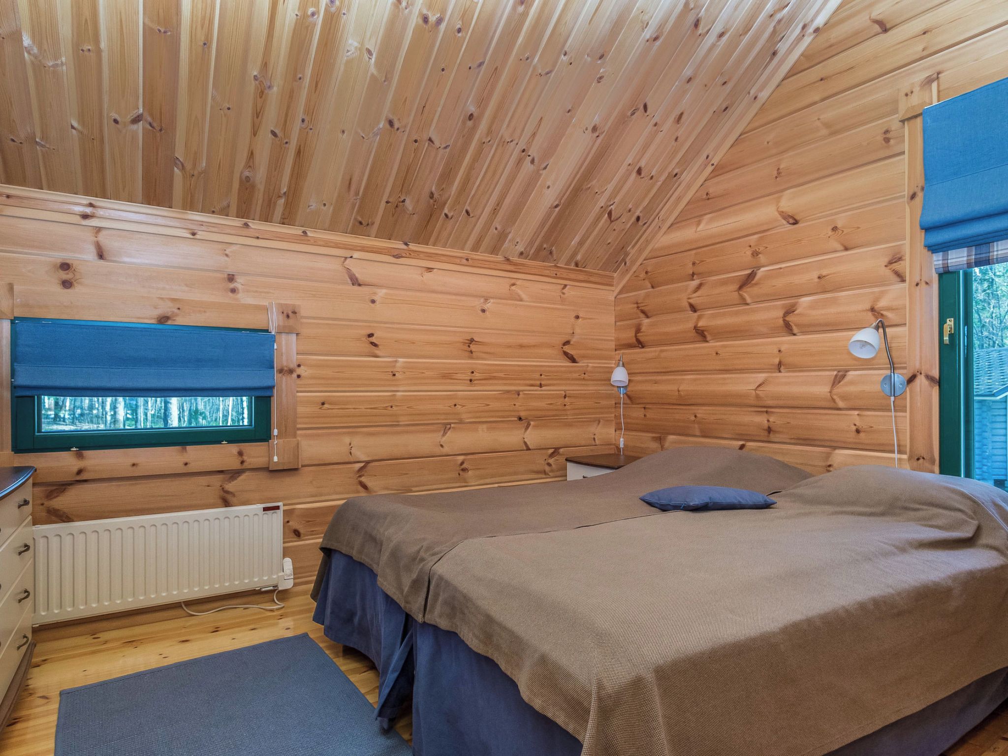 Un dormitorio de cabaña de madera con cama, lámparas de noche, ventana y radiador.