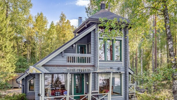 A two-story cabin with a balcony and a deck, surrounded by a forest.
