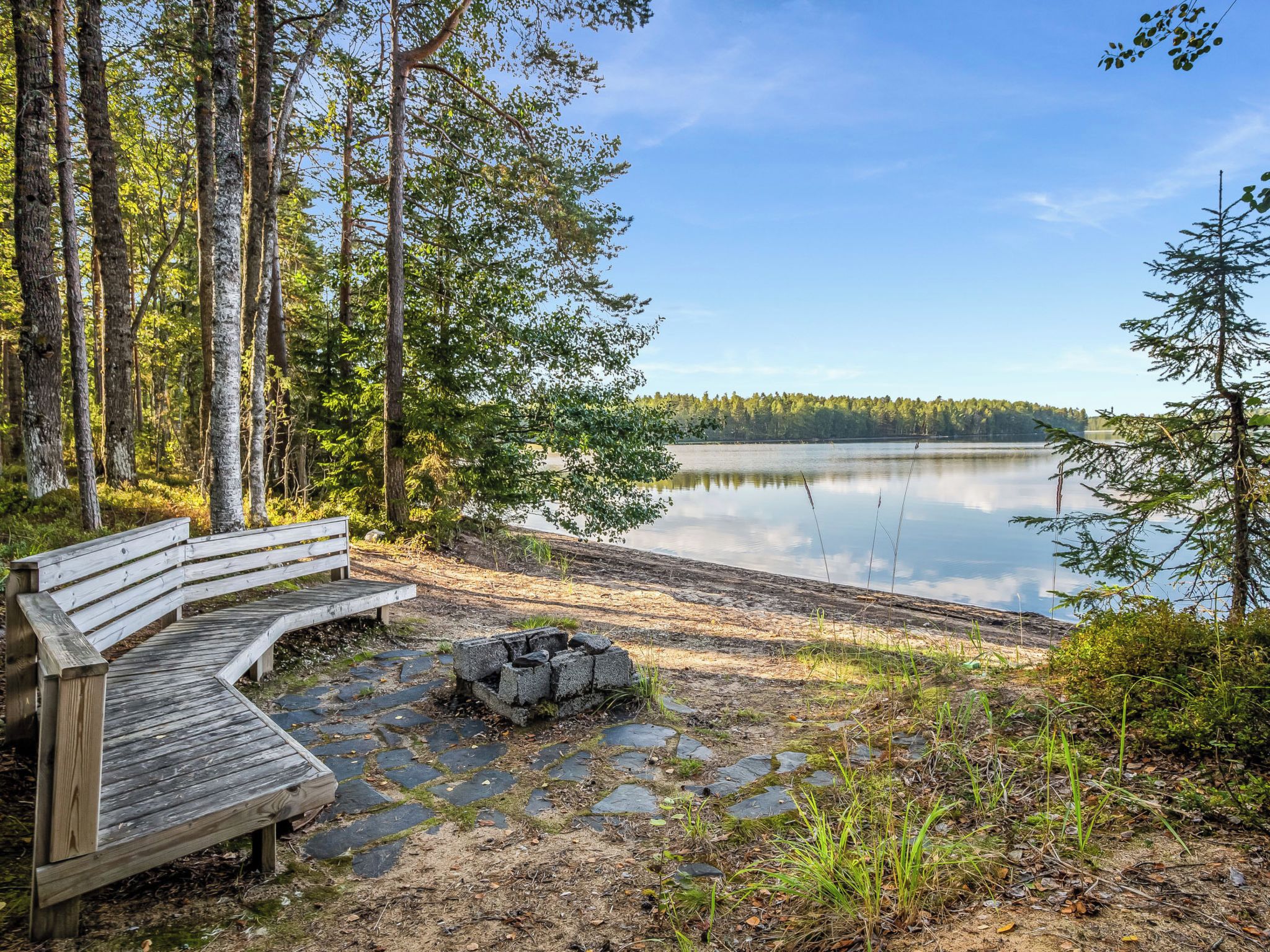 Puisesta penkistä ja kivestä rakennetusta nuotiopaikasta voi nauttia rauhallisen järven rannalla, jota ympäröivät puut.
