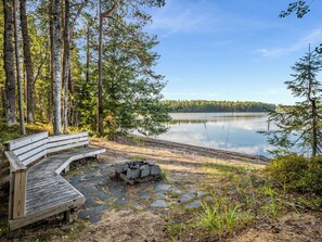 A wooden bench and a stone fire pit by a calm lake surrounded by trees. - Villa populus by Interhome (Pertunmaa)