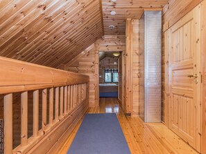 Un couloir intérieur en bois, avec une porte en métal, une rambarde en bois et un tapis bleu sur le sol.