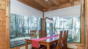 A wooden cabin with a dining table, chairs, and large windows overlooking a forest.
