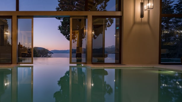 A modern indoor swimming pool with glass doors and windows offering a view of a mountain and lake at dusk. - Villa Beluno Hotel & Spa, small luxury hotels of the world (San Carlos de Bariloche)