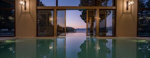 Piscina interior moderna con puertas y ventanas de vidrio que ofrecen vistas a una montaña y un lago al anochecer.