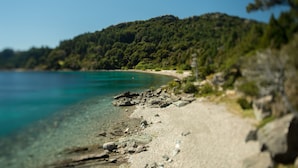 A beach with clear turquoise water, a rocky shoreline, and a forested hill in the background. - Villa Beluno Hotel & Spa, small luxury hotels of the world (San Carlos de Bariloche)