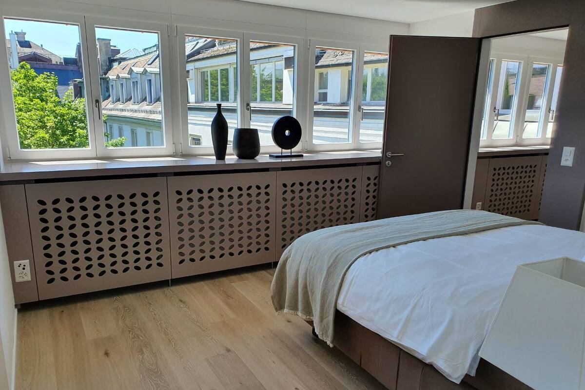Slaapkamer met een groot raam, een bed, een radiator en uitzicht op gebouwen.