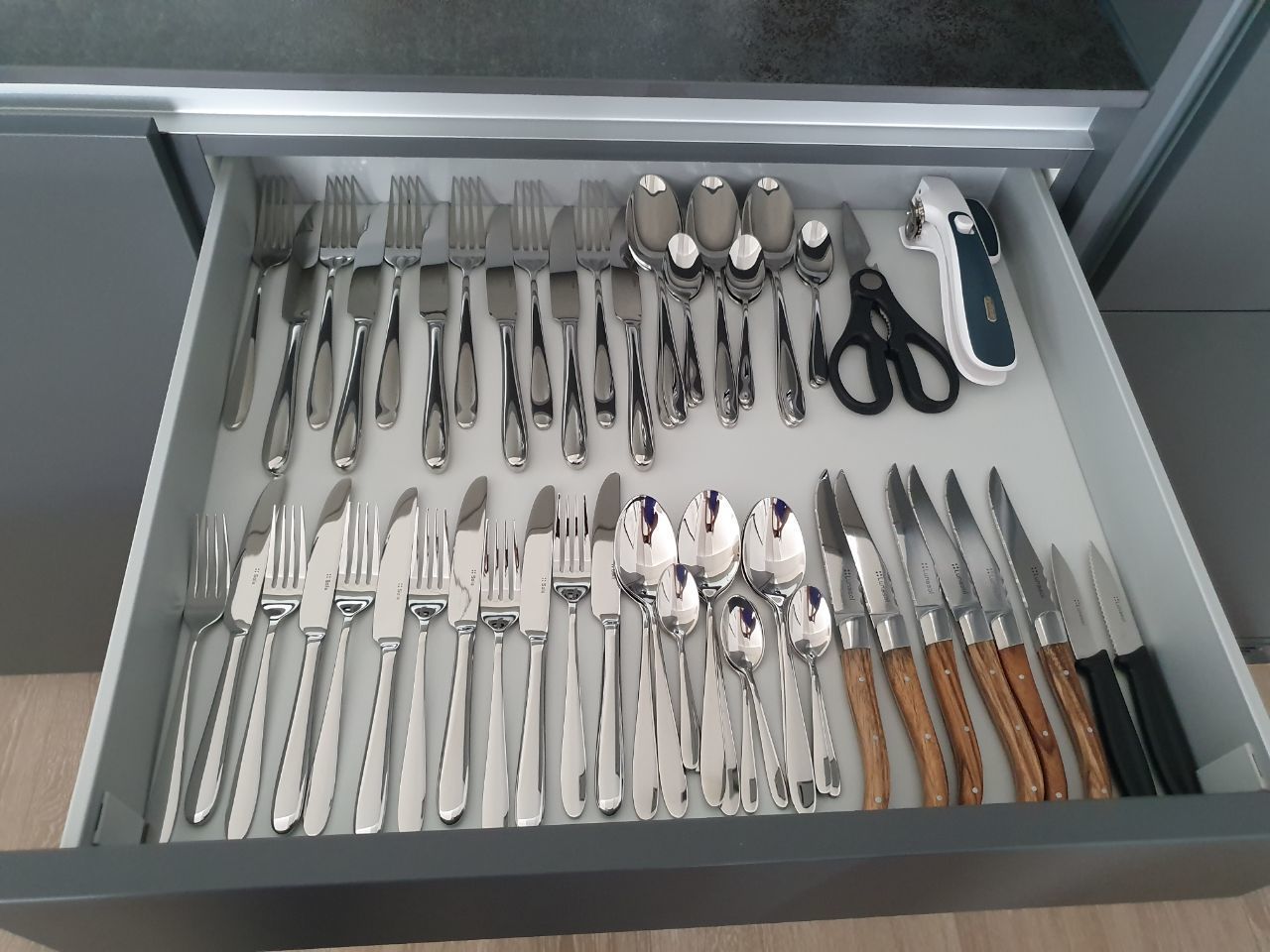 Eine Schublade mit verschiedenen Besteckstücken, darunter Gabeln, Messer und Löffel, sowie eine Schere.