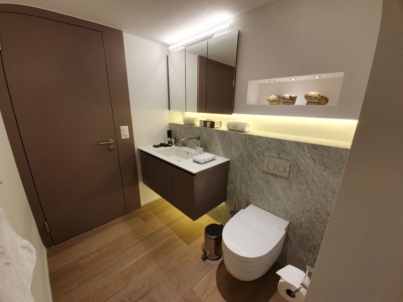 A modern bathroom with a wooden floor, a white toilet, a sink with a mirror above, and a cabinet with a towel on top.