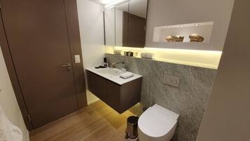 A modern bathroom with a wooden floor, a white toilet, a sink with a mirror above, and a cabinet with a towel on top.