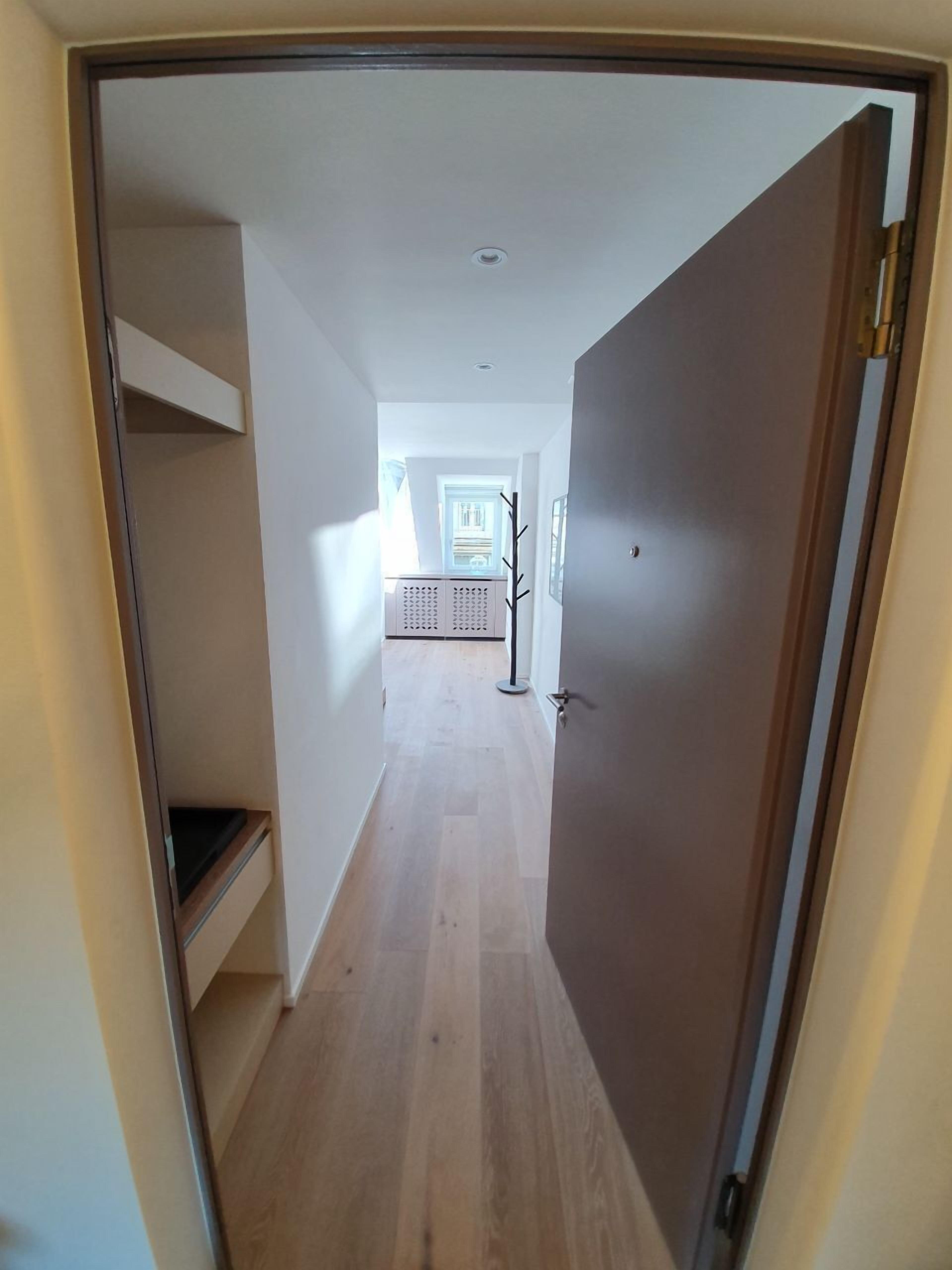 A narrow hallway with a wooden floor, a door on the right, and a view into another room with a window and a radiator.