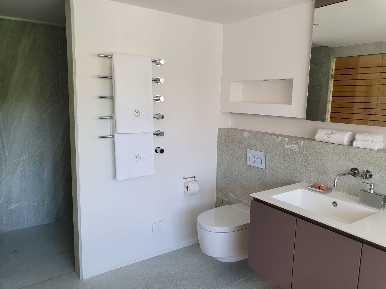 Ein modernes Badezimmer mit einer weißen Toilette, einem Waschbecken mit Spiegel und einem Handtuchhalter mit mehreren Handtüchern.