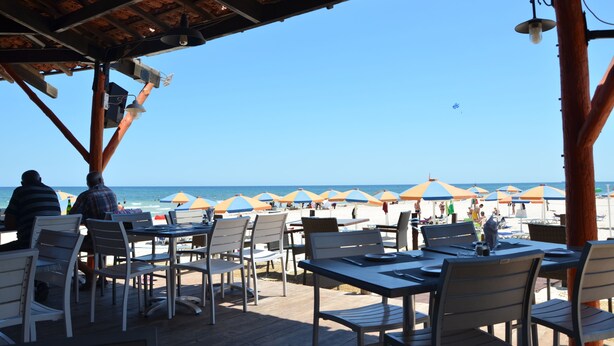 Een terras met tafels en stoelen met uitzicht op een strand met parasols.