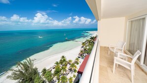 A balcony with a view of a beach and ocean, featuring white chairs and a palm tree-lined walkway.
