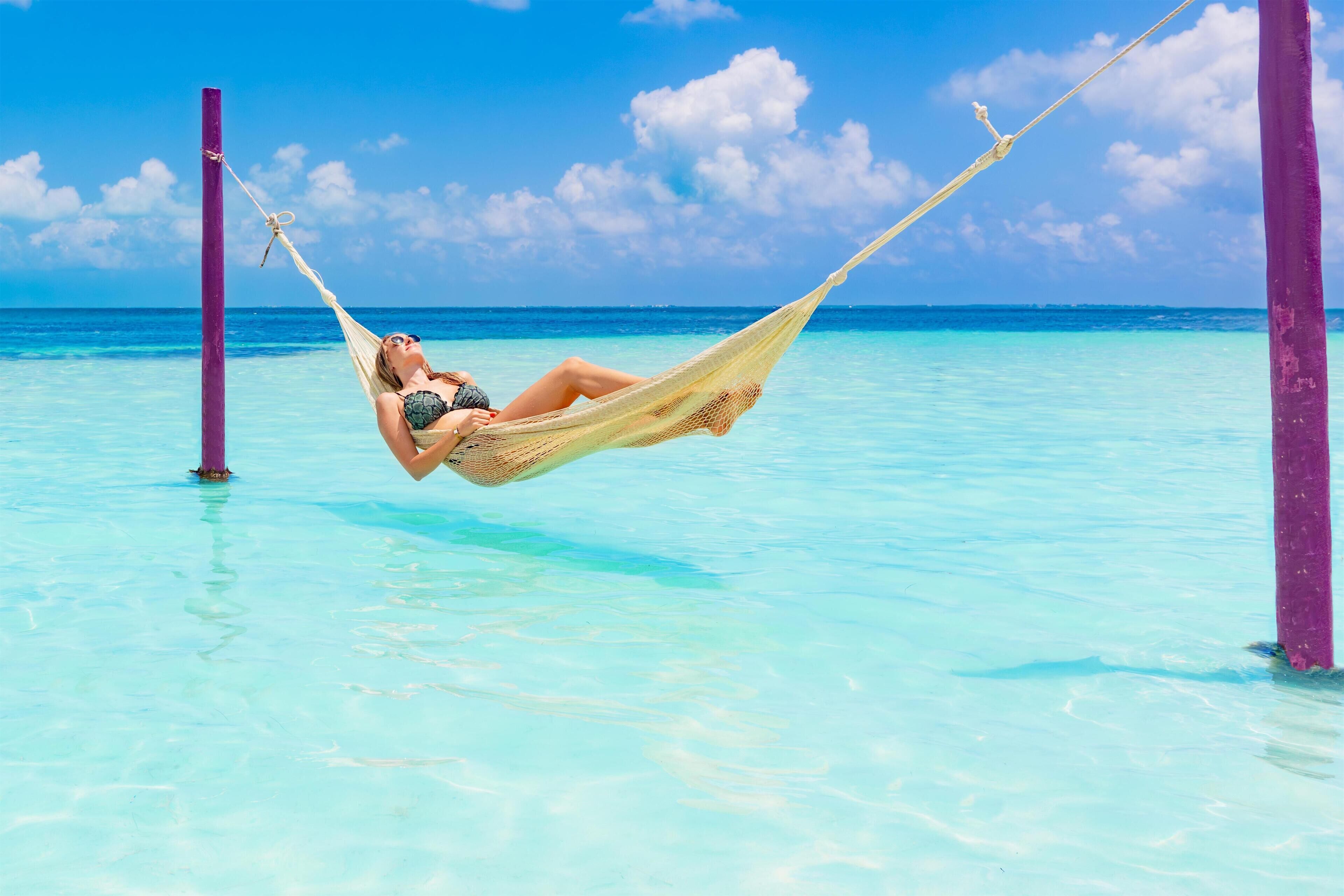 A person relaxing in a hammock between two purple poles in clear turquoise water.