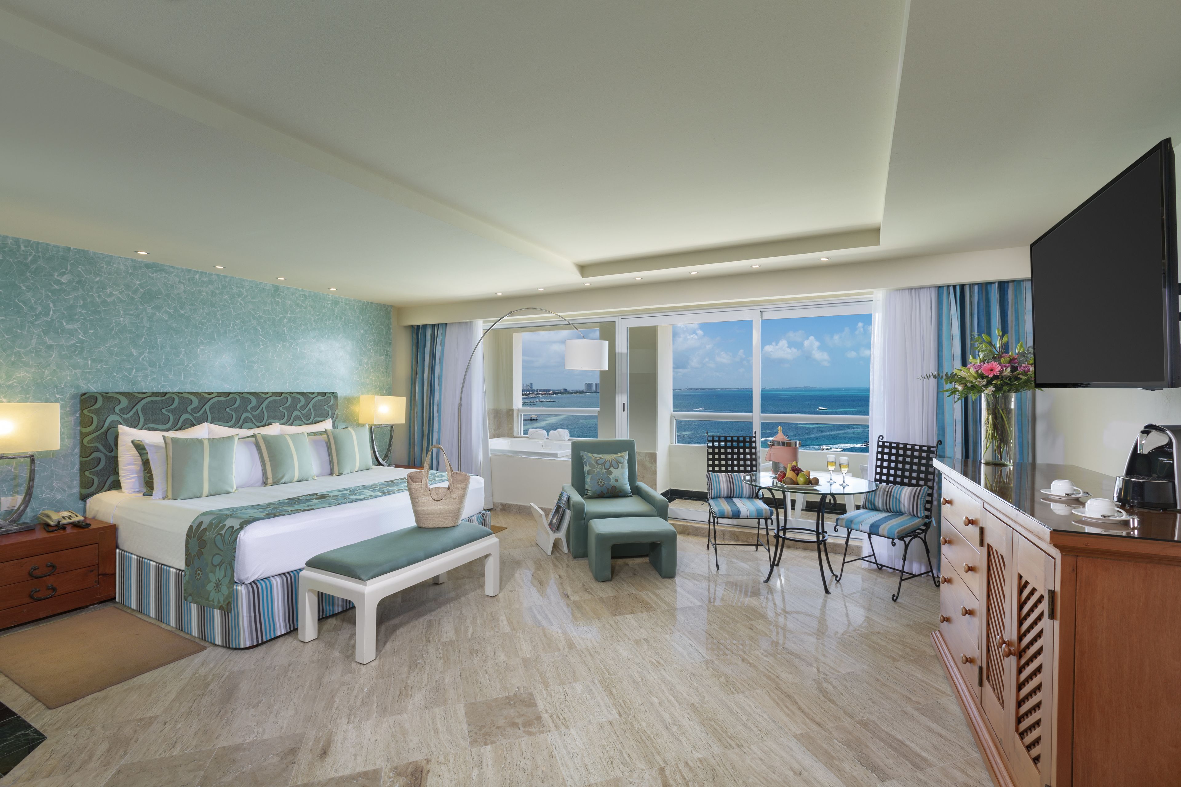 Une chambre d’hôtel avec un grand lit, un coin salon avec une table et des chaises, une vue sur l’océan et un téléviseur fixé au mur.