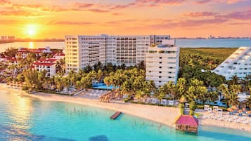 Un hotel frente a la playa con varias edificaciones, una piscina y zona de playa con sombrillas y sillas de descanso.