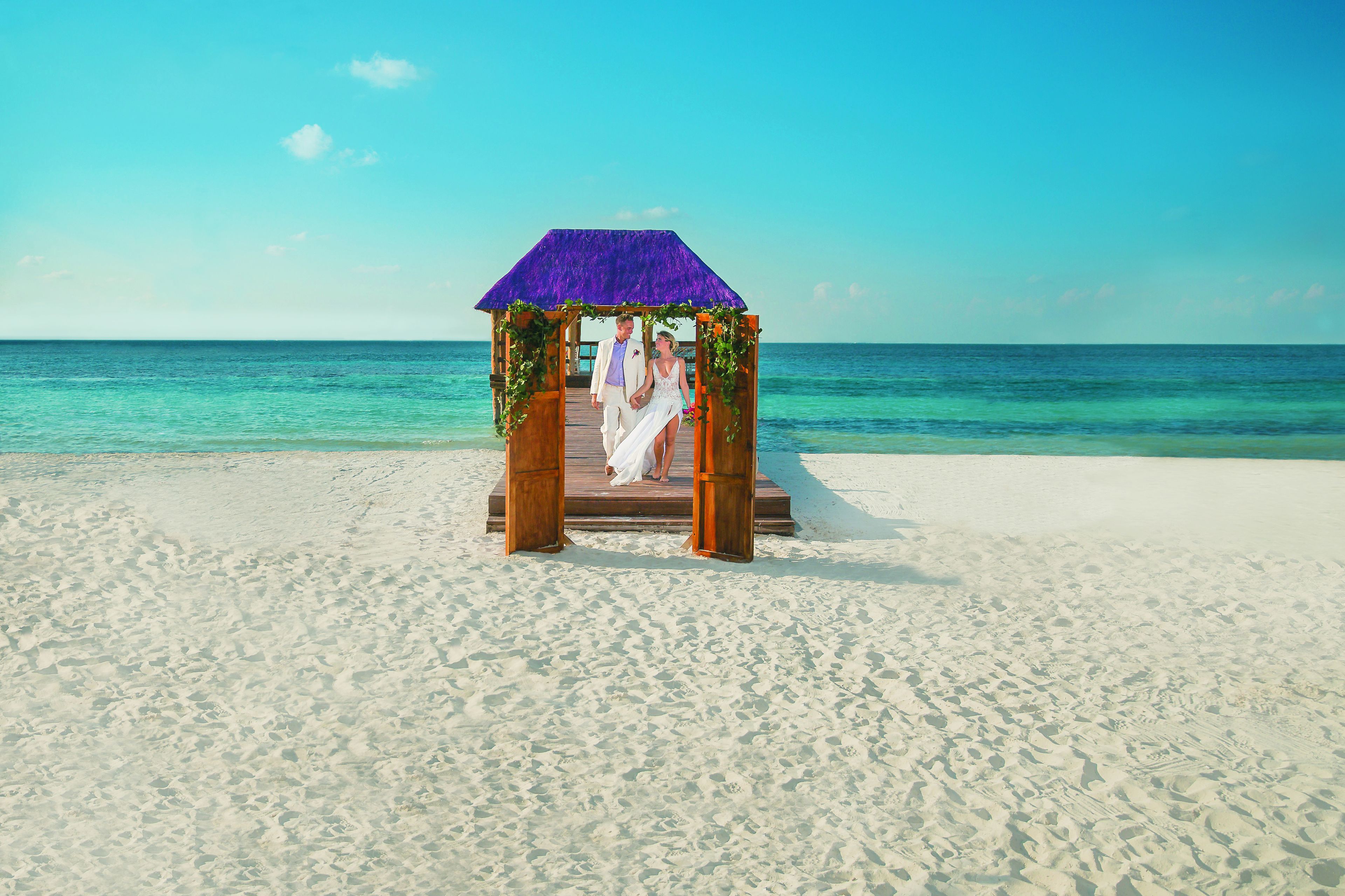 Um arco de casamento na praia, com estrutura de madeira e teto roxo, montado sobre areia, com águas azuis cristalinas ao fundo.