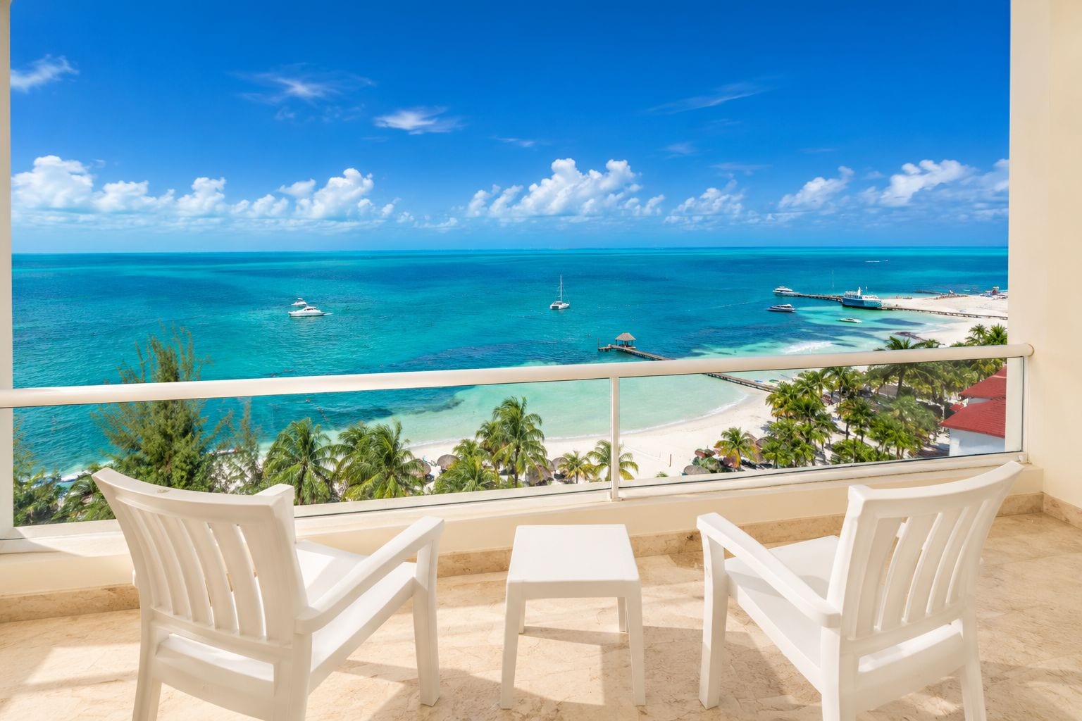 A balcony with two white chairs and a small table, overlooking a turquoise sea with boats and a beach.