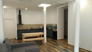 A modern living room with a gray sectional sofa and a wooden bench, a kitchen with dark cabinets and a black range hood, and a patterned wooden floor. - Hub Hostel Seville (Seville)