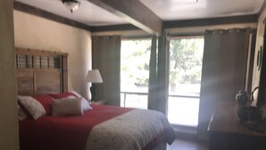 A bedroom with a large bed, a nightstand, a lamp, and a view of the outdoors through a window. - North Wind Condo (Angel Fire)