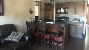 A living room with a dark leather sofa and a dining area with wooden chairs and a table. A kitchen with wooden cabinets and a microwave is visible in the background. - North Wind Condo (Angel Fire)