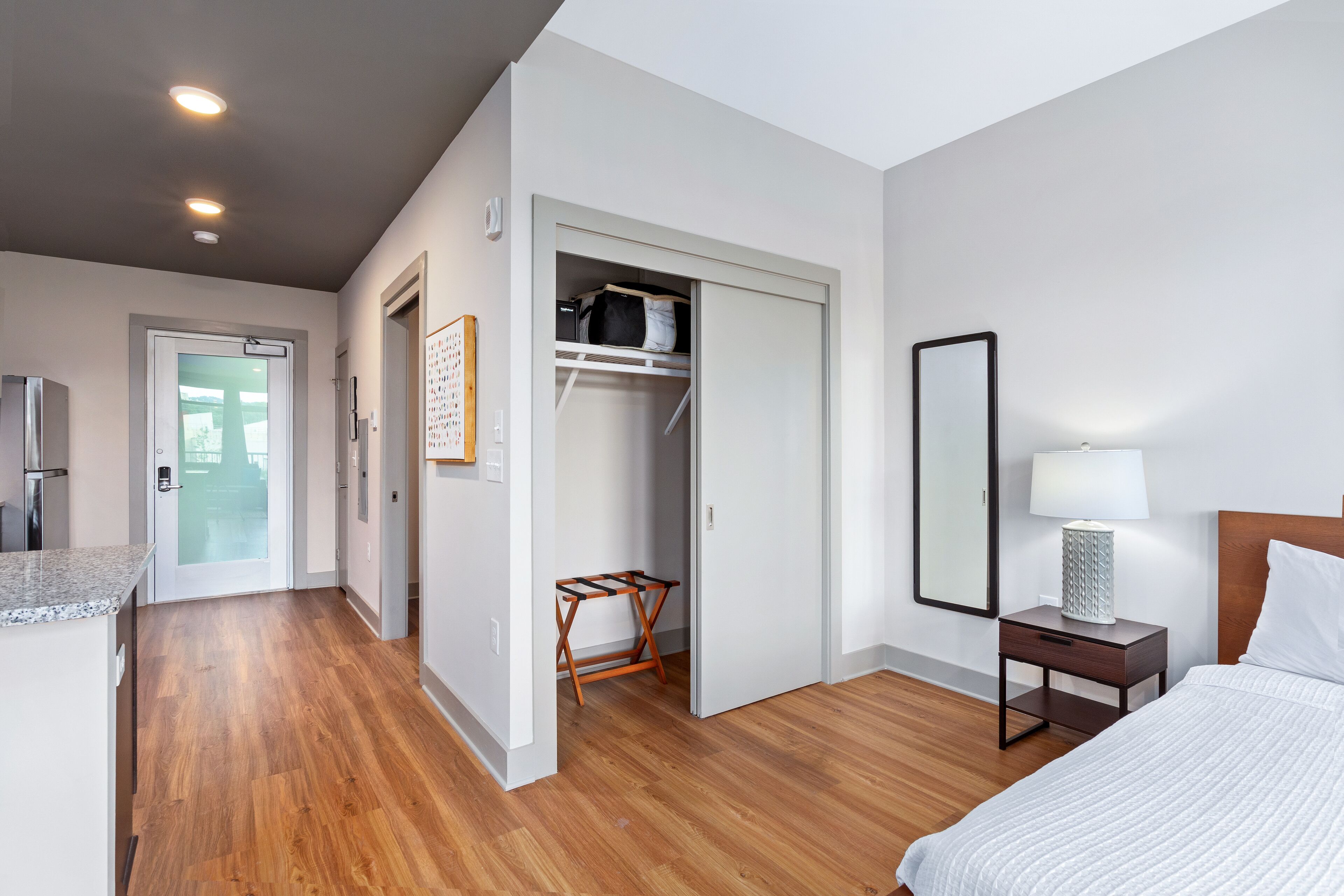 A hotel room with a wooden floor, a bed with white bedding, a nightstand with a lamp, a wardrobe, and a door leading to the outside.