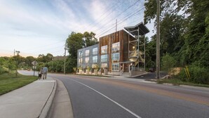 A modern multi-story building with a mix of wood and metal exterior, a visible entrance, and a sidewalk adjacent to the road. - River Row Flats (Asheville)
